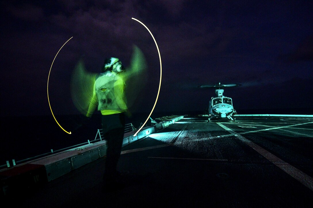 A sailor directs a helicopter at night on a ship's deck in the ocean.