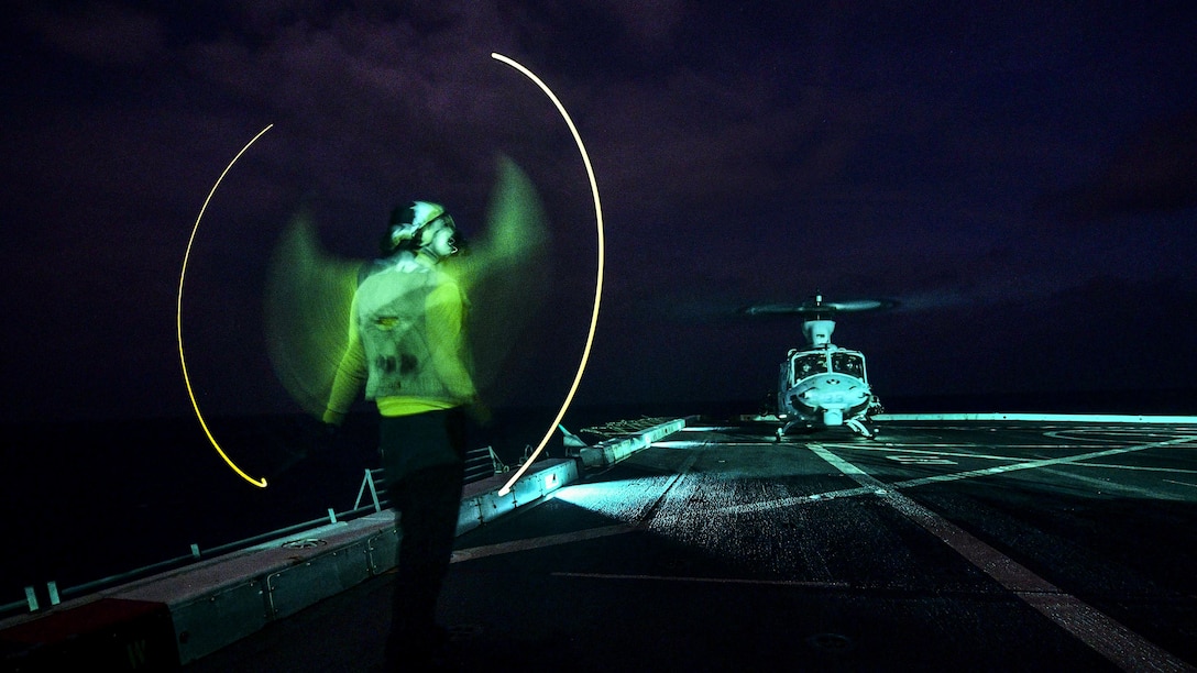 A sailor directs a helicopter at night on a ship's deck in the ocean.