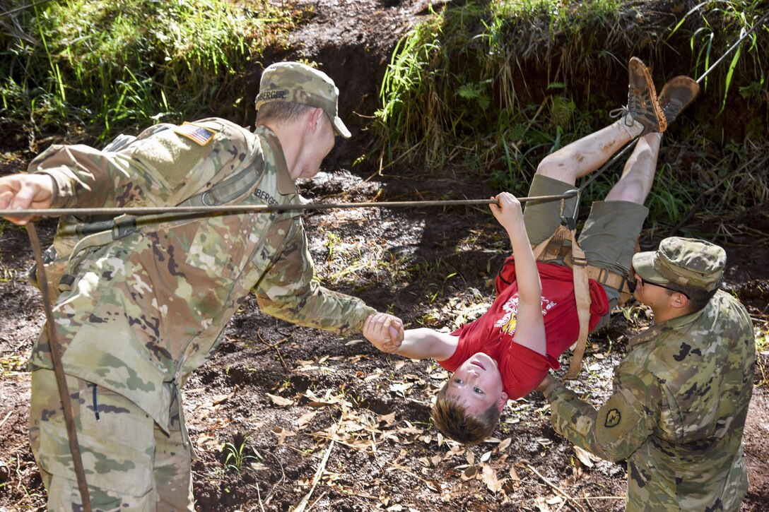 Two soldiers help a Cub Scout crawl upside down over a rope line.