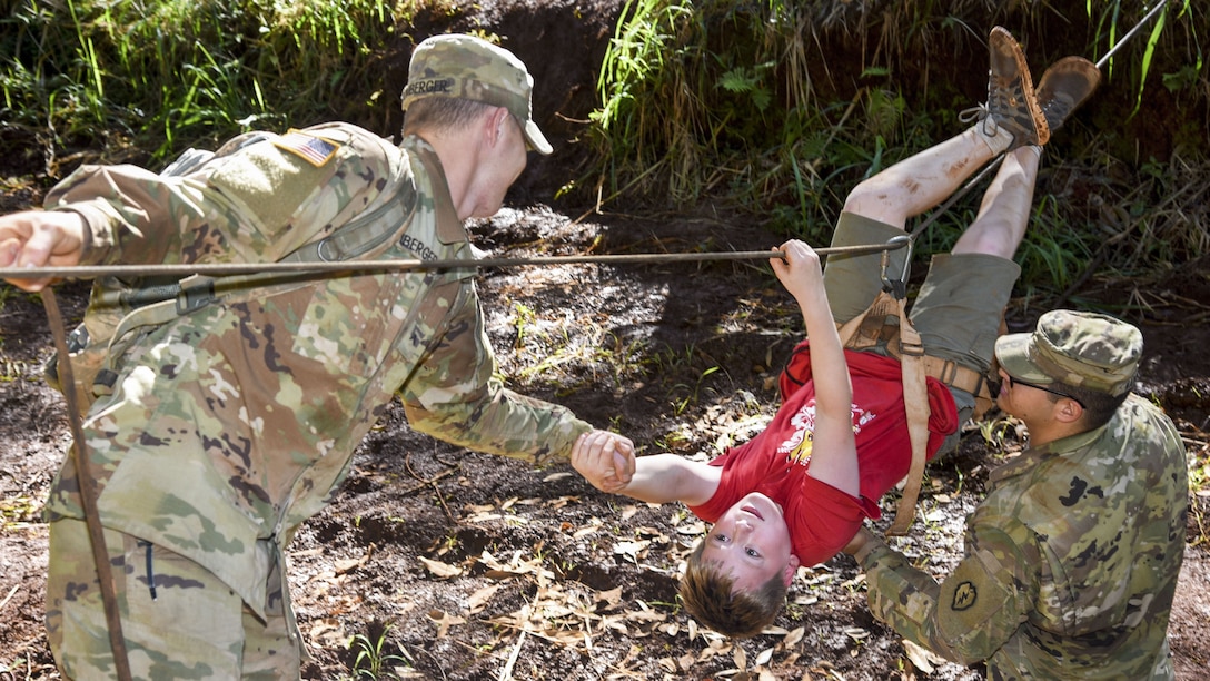 Two soldiers help a Cub Scout crawl upside down over a rope line.