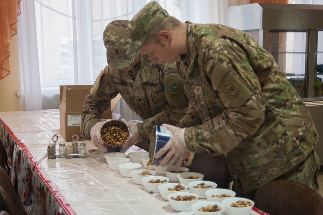 Two soldiers pour food into two rows of bowls on a table.