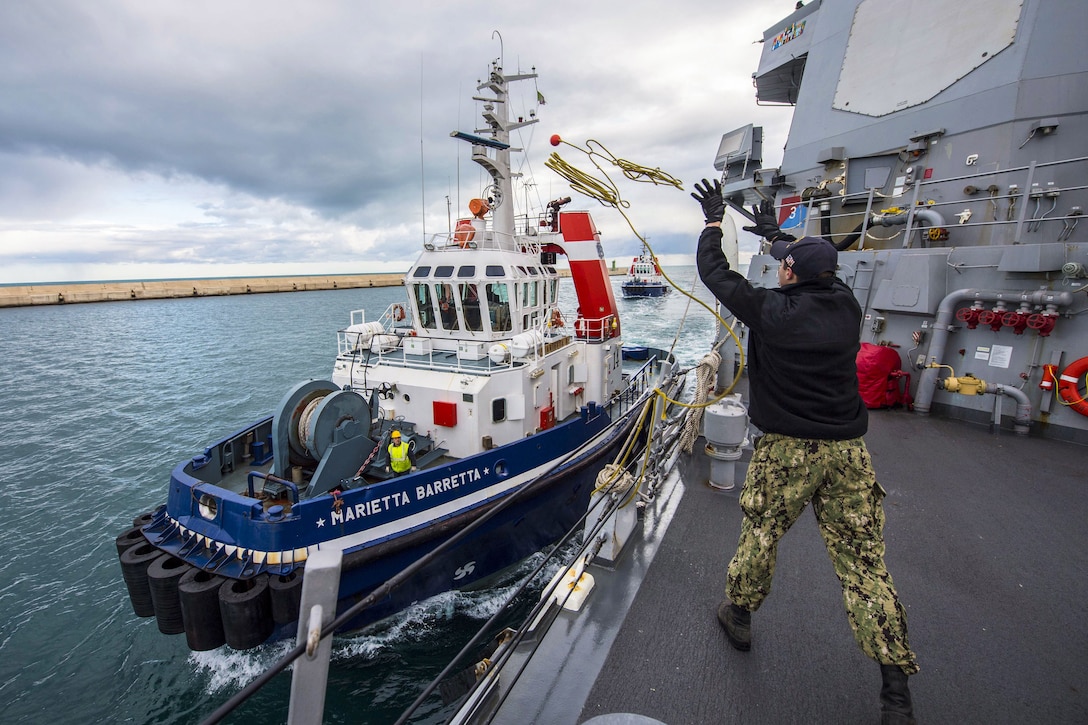 A sailor on a ship tosses a line to a tugboat.