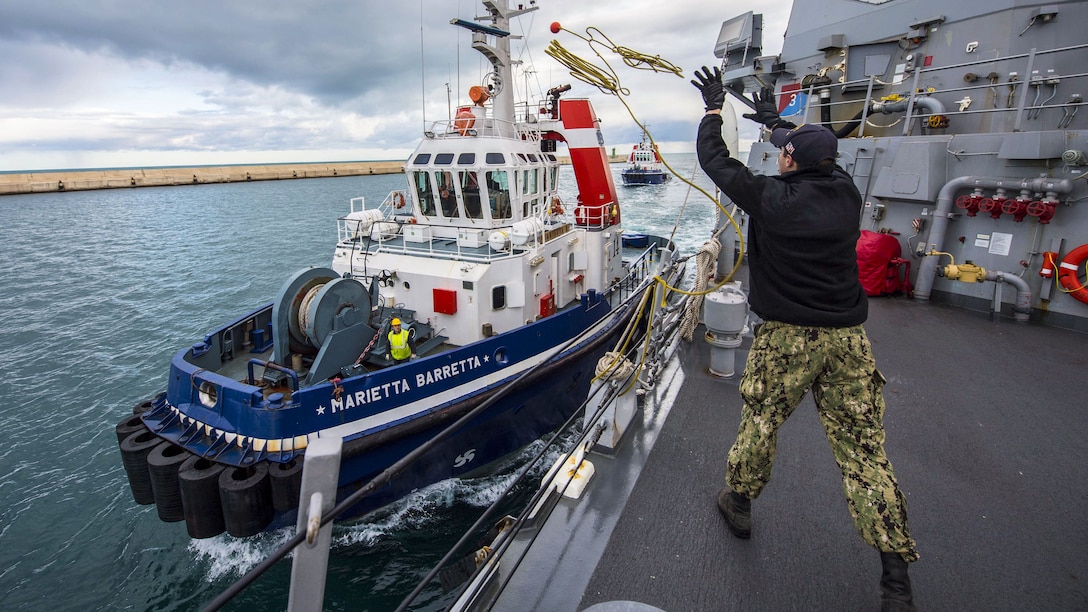 A sailor on a ship tosses a line to a tugboat.