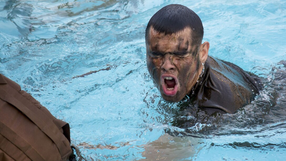 A Marine wearing face paint a tan shirt takes a breath while swimming in a pool.