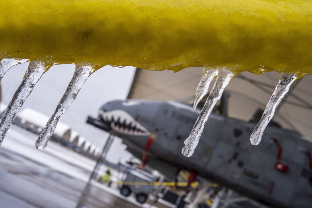 Icicles hang from a yellow structure on a flight line, as an aircraft with a scowling face painted on it sits in the background.