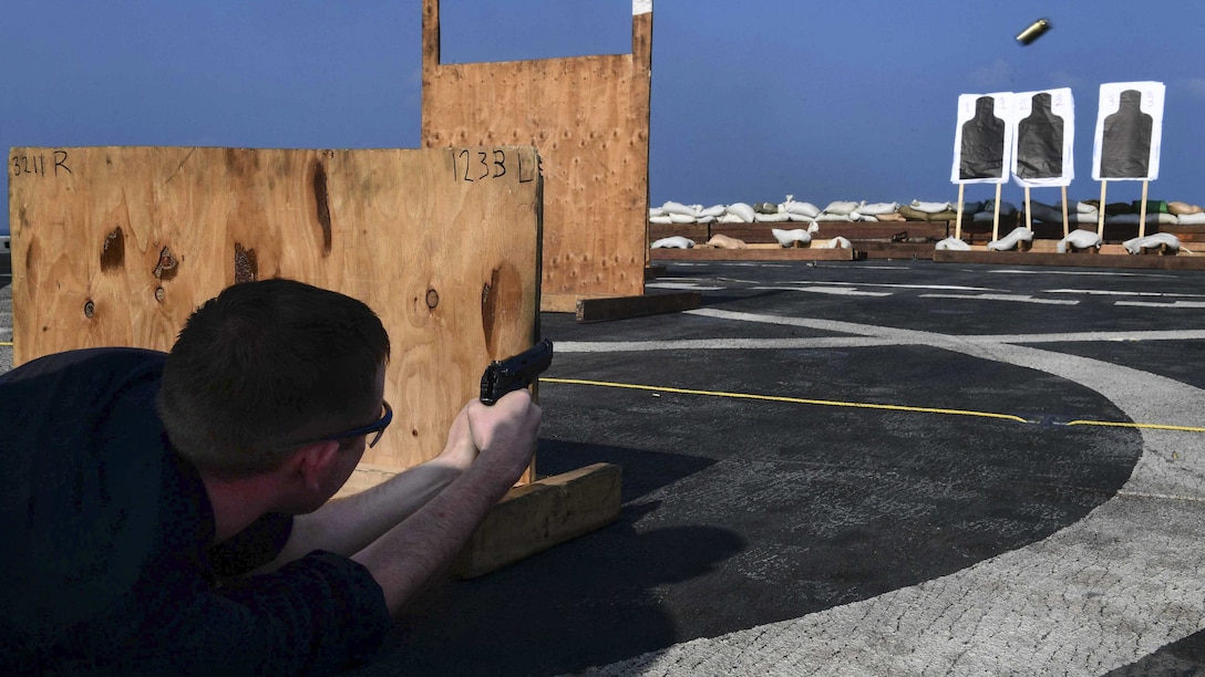 A sailor lies on a flight deck and fires a pistol at targets while behind a wooden barrier.