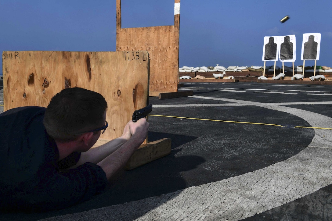 A sailor lies on a flight deck and fires a pistol at targets while behind a wooden barrier.