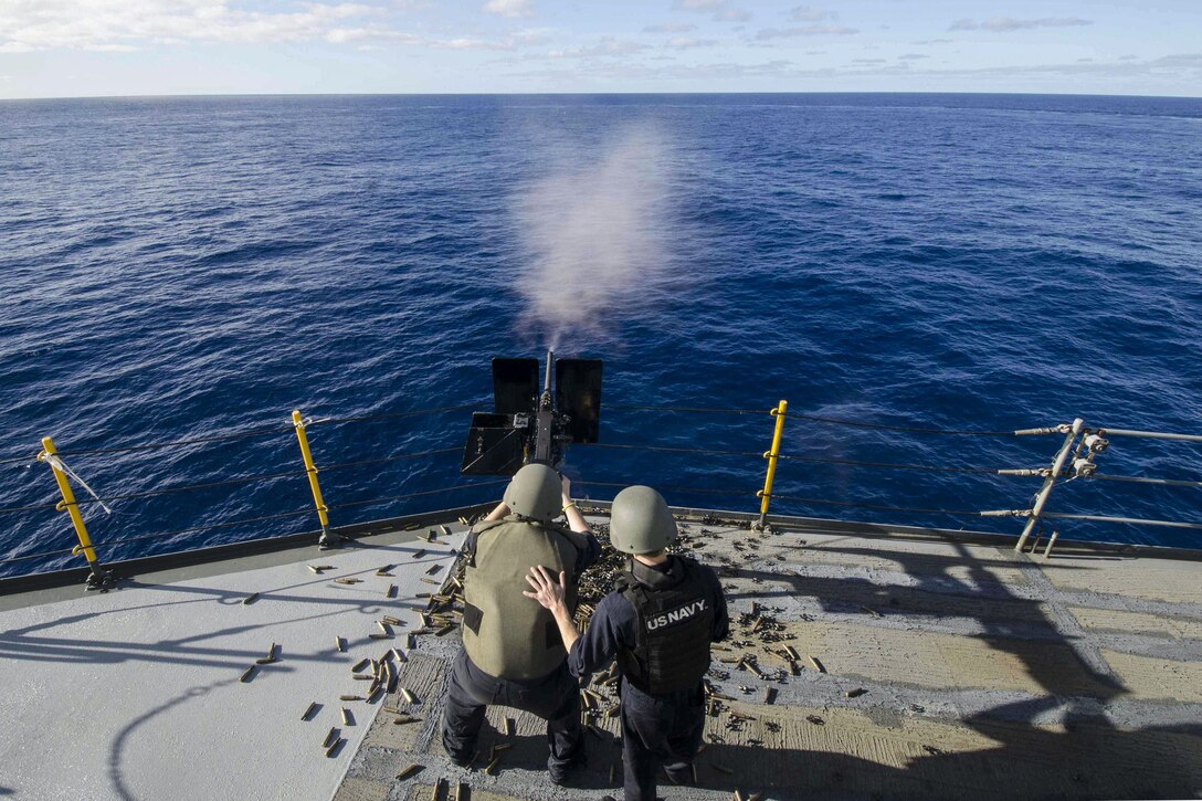 A sailor fires a machine gun mounted on a ship toward the sea, as another sailor places his hand on the first sailor's back.