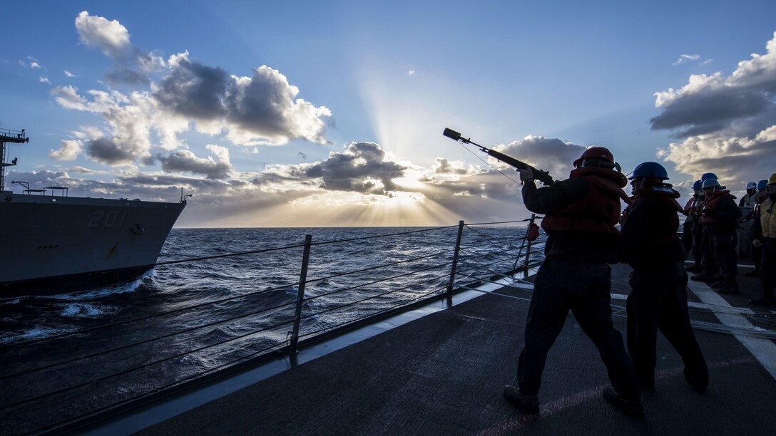 A sailor, shown in silhouette, aims a shot-line gun on a ship's deck toward another ship nearby.