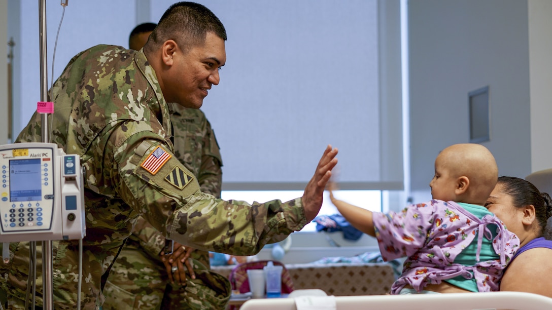A soldier high-fives an infant sitting on a woman's lap in a hospital room.