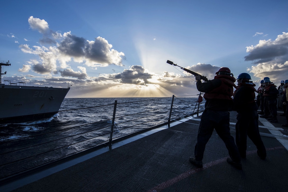 A sailor, shown in silhouette, aims a shot-line gun on a ship's deck toward another ship nearby.