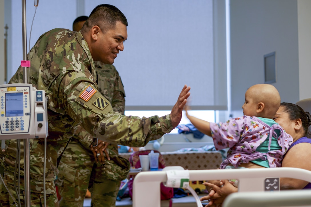 A soldier high-fives an infant sitting on a woman's lap in a hospital room.