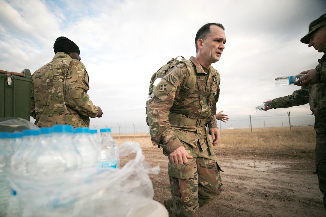 A soldier walks past other soldiers and bottles of water on a table.