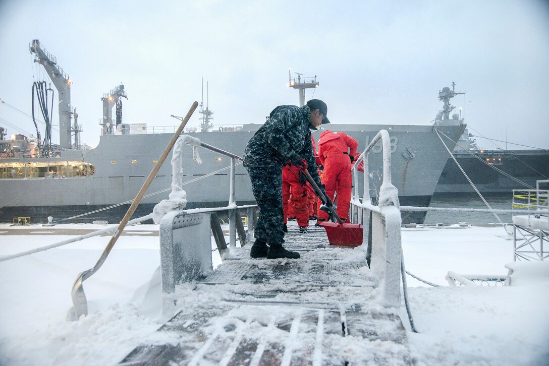 Sailors shovel snow off the walkway to a ship.
