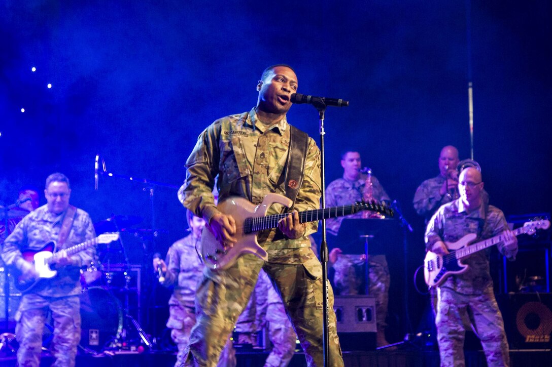 A soldier plays guitar and sings as fellow soldiers play guitar behind him on a blue-lit, smoky stage.