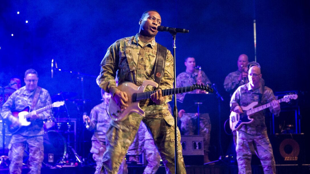 A soldier plays guitar and sings as fellow soldiers play guitar behind him on a blue-lit, smoky stage.