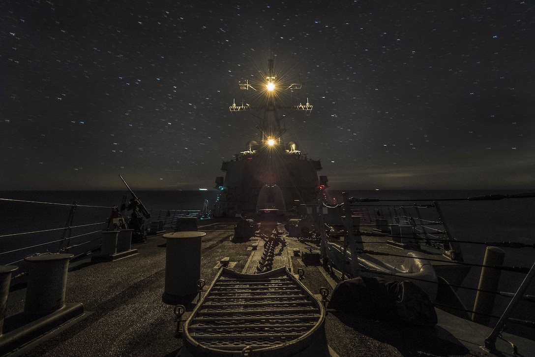 Lights illuminate the top of a ship as it travels, against the backdrop of a starry night sky.