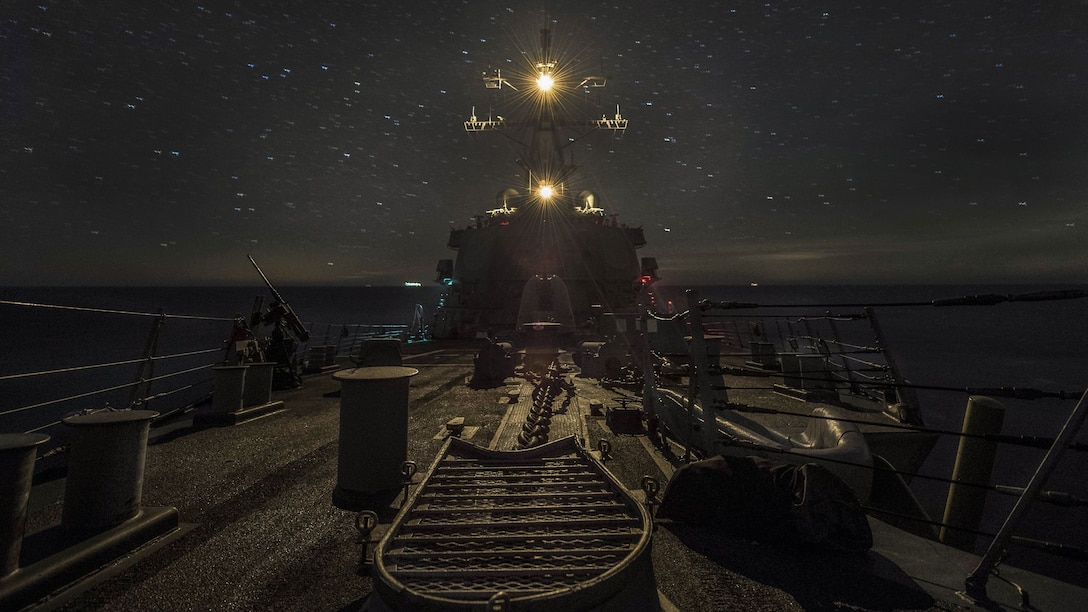 Lights illuminate the top of a ship as it travels, against the backdrop of a starry night sky.