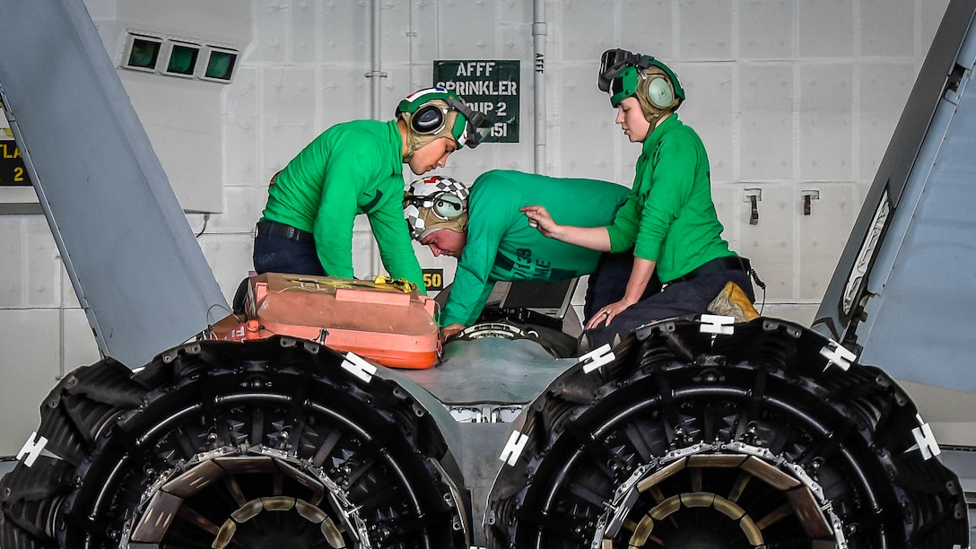 Three sailors in green shirts and helmets kneel on top of an aircraft, shown from behind.