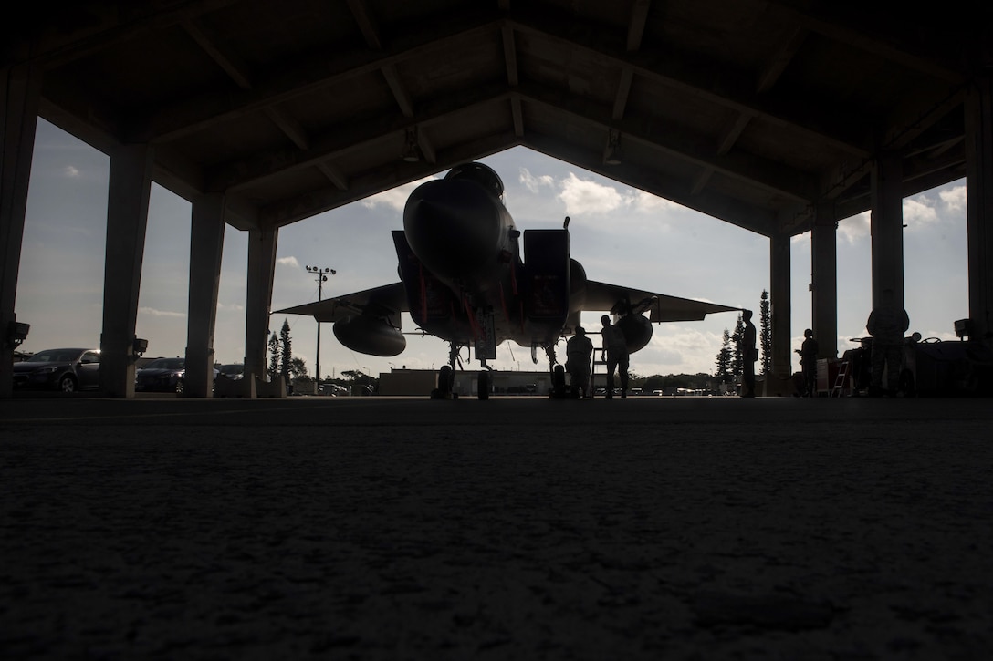 Airmen, shown in silhouette, work on a fighter jet in a hangar with a jagged roof line.
