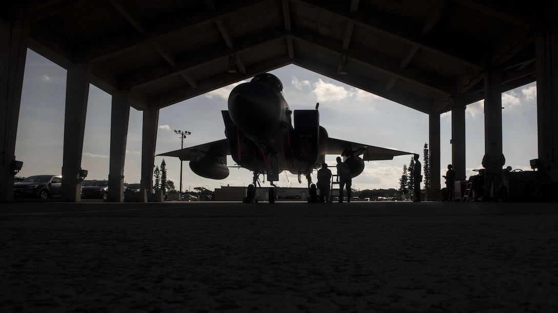 Airmen, shown in silhouette, work on a fighter jet in a hangar with a jagged roof line.