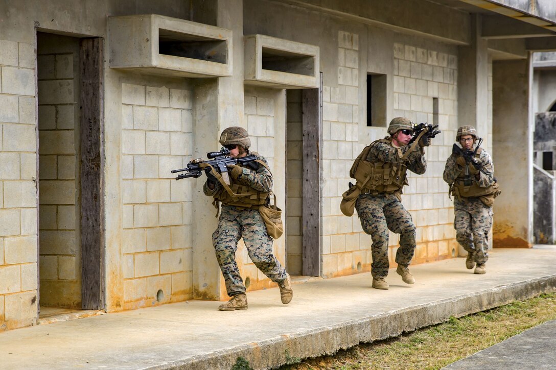 Three Marines point rifles in different directions while walking in a line along an outside wall of a building.