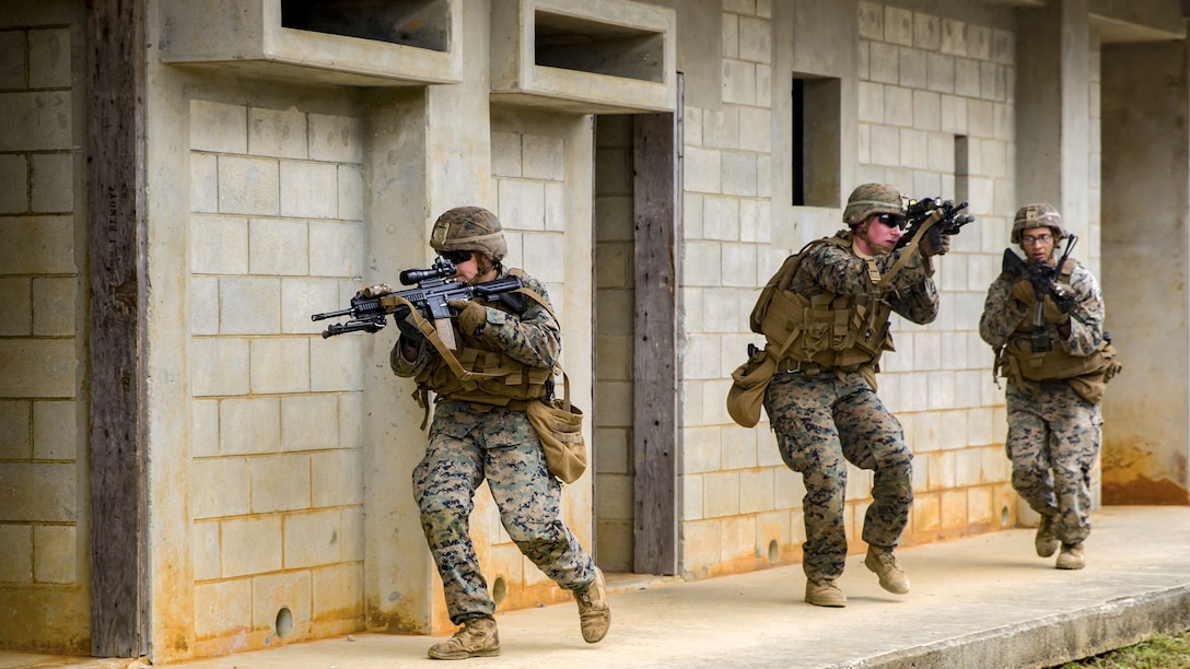 Three Marines point rifles in different directions while walking in a line along an outside wall of a building.