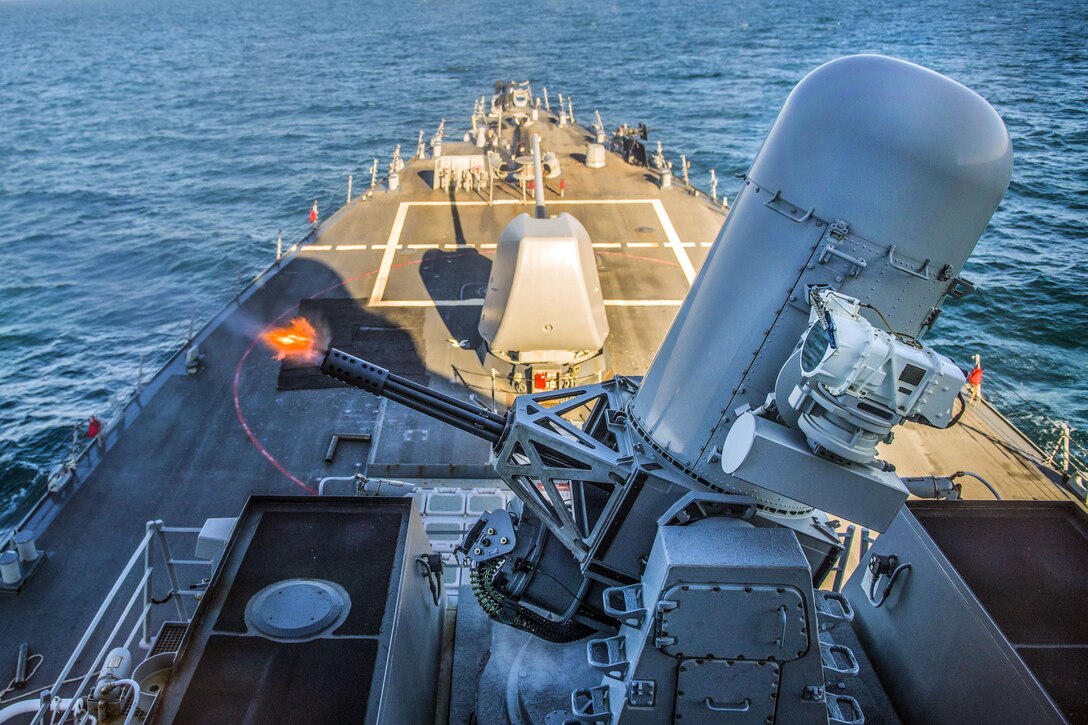 Flames burst from a large weapon system aboard a ship traveling in the sea.