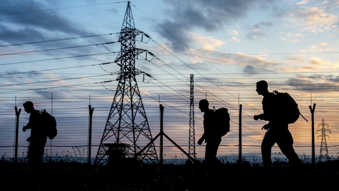 Three soldiers, shown in silhouette against a blue and pink sky, walk past an electrical tower.