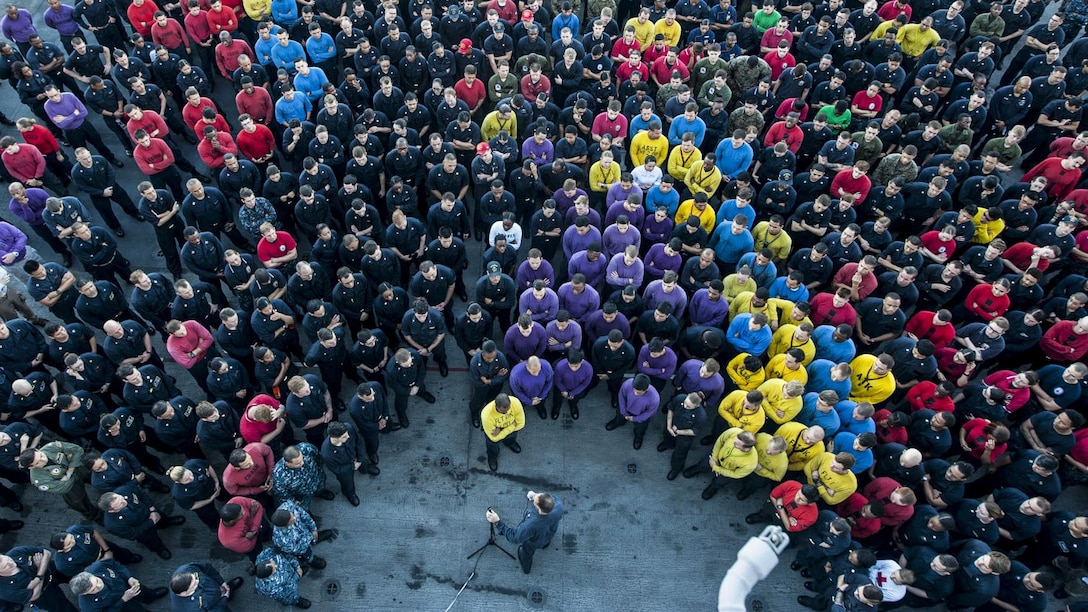 Dozens of sailors wearing different colors, shown from overhead, stand in a U-shape around an officer.