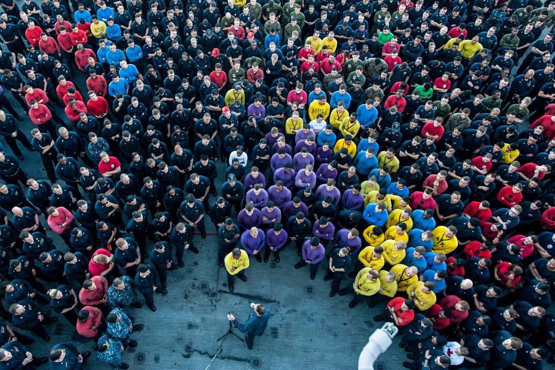 Dozens of sailors wearing different colors, shown from overhead, stand in a U-shape around an officer.