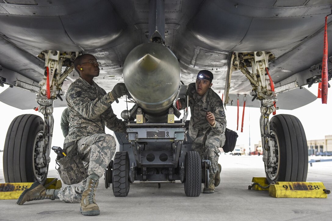 An airman kneels and another crouches while mounting a bomb on the underside of an aircraft.