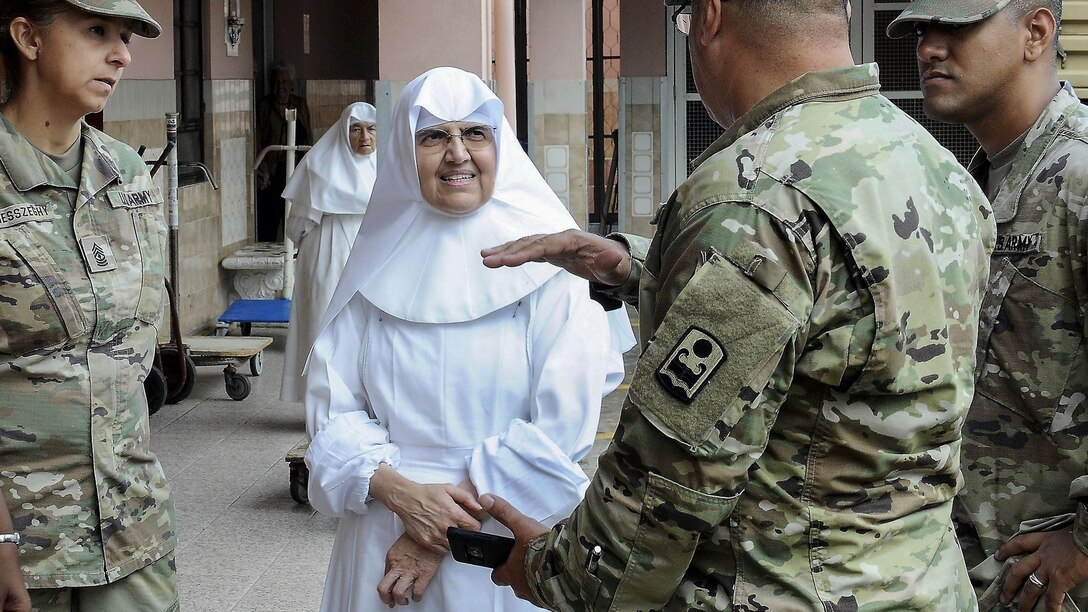 A soldier gesticulates while speaking to a nun and fellow soldiers outside a building.