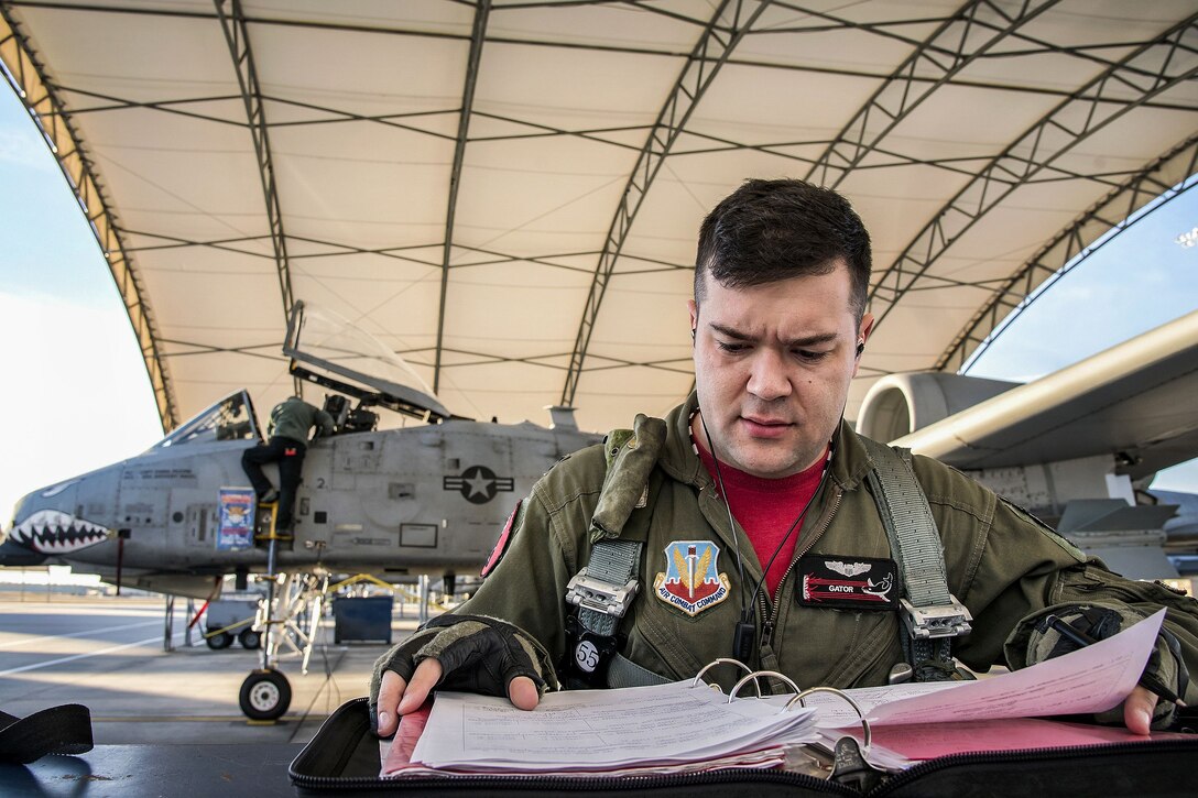 A pilot looks at a notebook on a flightline as personnel work on a fighter jet in the background.