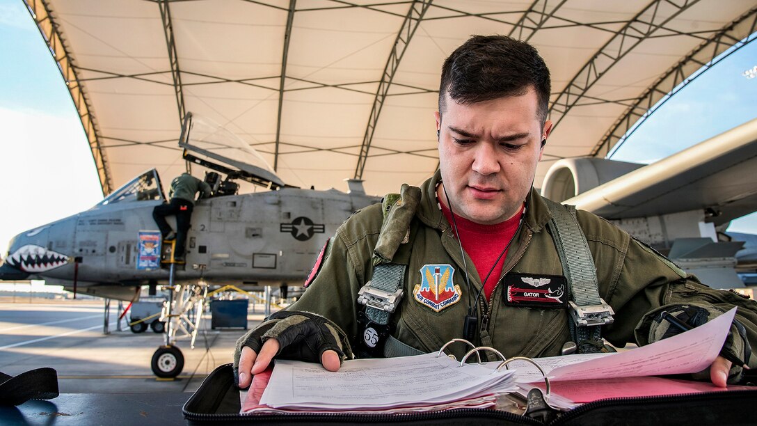 A pilot looks at a notebook on a flightline as personnel work on a fighter jet in the background.