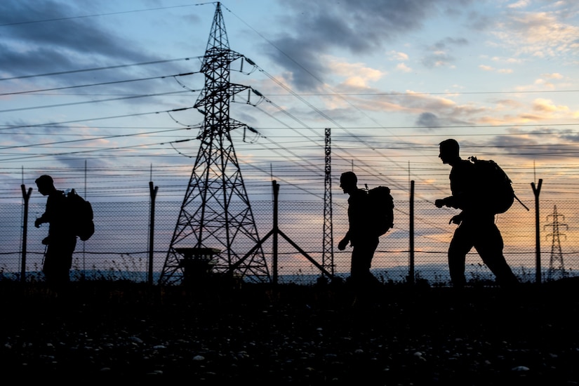 Three soldiers, shown in silhouette against a blue and pink sky, walk past an electrical tower.