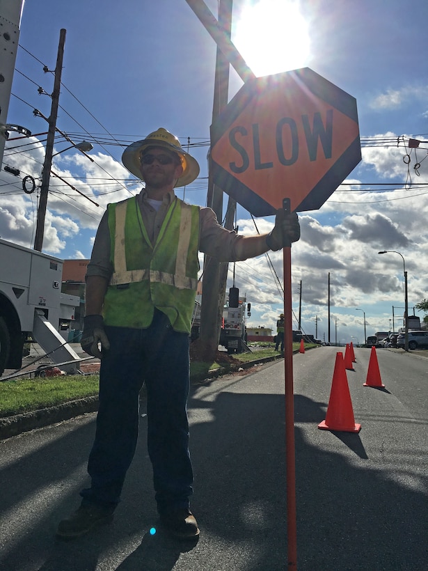 A flagman signals traffic to slow as they pass a crew of linemen from Fluor subcontractor MasTec working on a distribution line in San Juan, Puerto Rico, Dec. 12, 2017, as part of Task Force Power Restoration's mission to repair the island's damaged electrical grid.