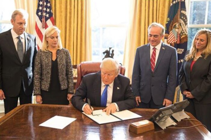 President Donald J. Trump signs a document at his desk as officials stand around him.