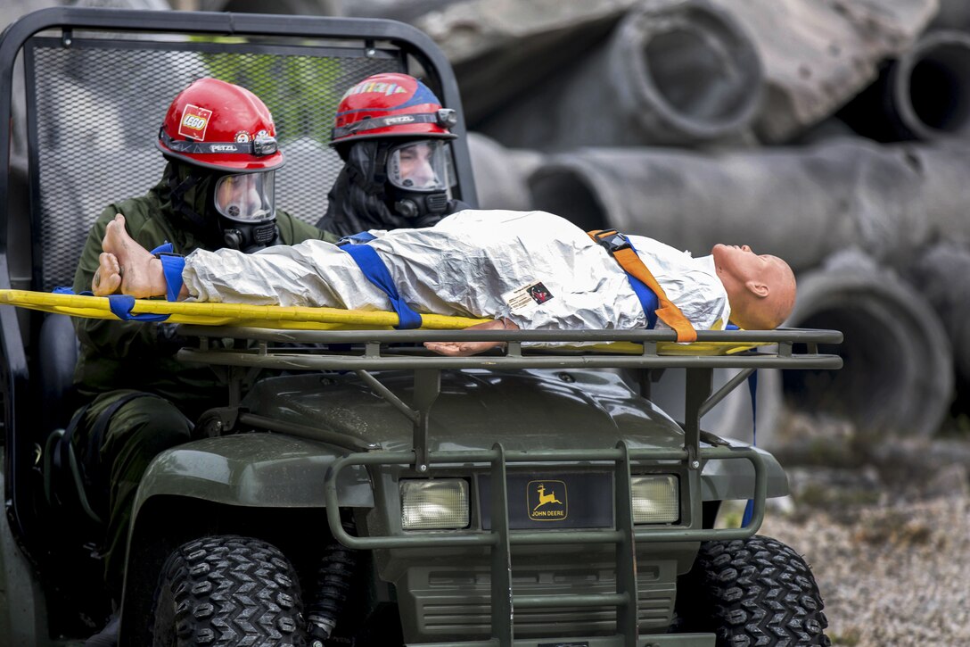 Soldiers wearing red helmets drive a jeep with a dummy on a litter strapped to the front bumper.