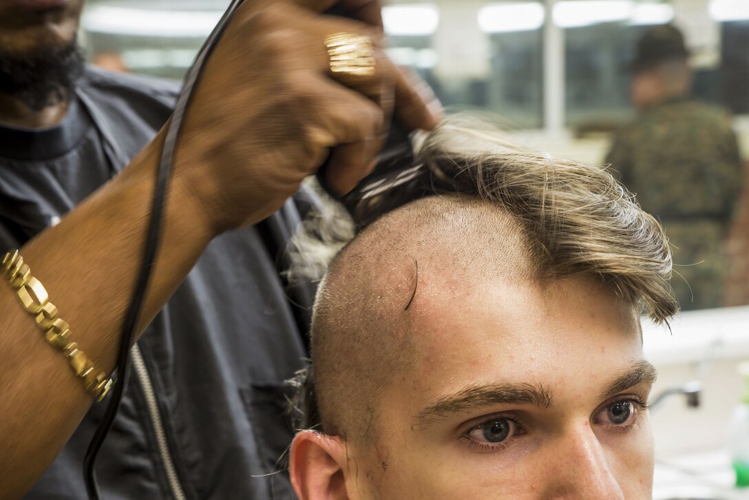 A recruit stares ahead as someone continues to shave his head, which is half-shorn.