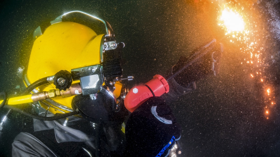 Fire and sparks shoot from a torch held by a sailor in a yellow diving helmet.