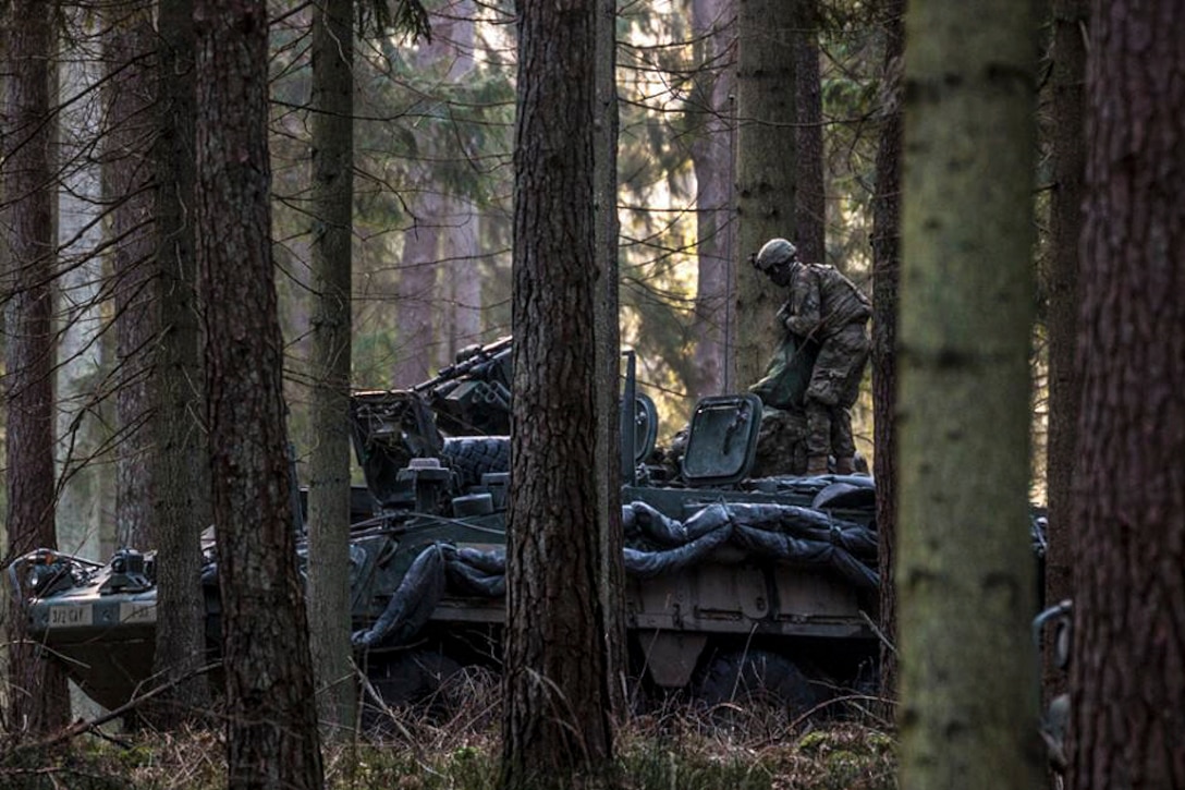 A service member stands atop a tank in a forested area.