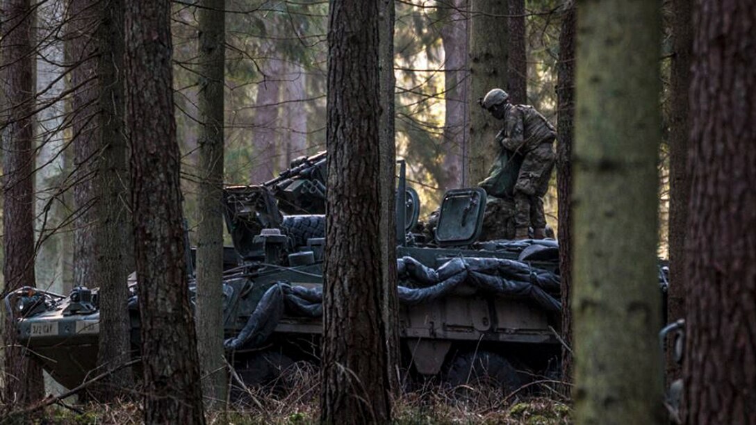 A service member stands atop a tank in a forested area.