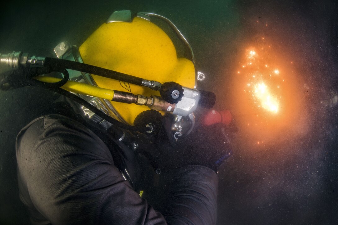 Fire and sparks shoot from a torch held by a sailor in a yellow diving helmet.