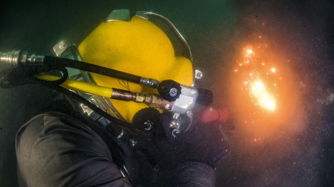 Fire and sparks shoot from a torch held by a sailor in a yellow diving helmet.