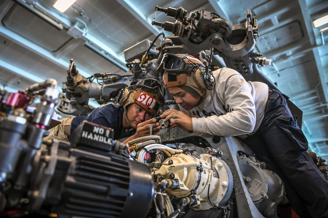 Two sailors perched atop a helicopter look at the back of a digital camera.