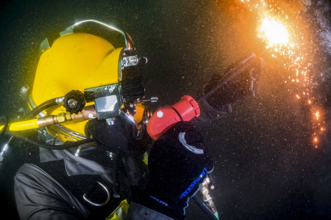 Fire and sparks shoot from a torch held by a sailor in a yellow diving helmet.