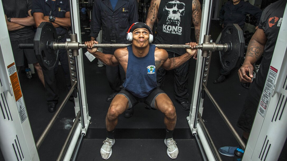 A sailor squats while holding a barbell on his shoulders as others observe.