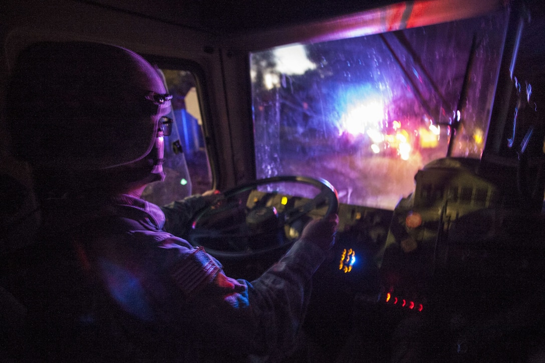 A soldier wearing a helmet drives a tactical vehicle in the dark, as colored lights illuminate the windshield.