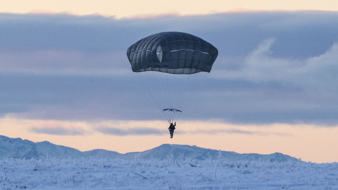 A soldier parachutes above a backdrop of snowy mountains and pale blue and pink sky.